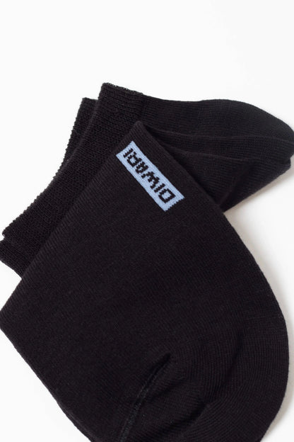 A pair of black ankle socks folded against a white background, featuring a light blue rectangular label with dark blue text on the top cuff.