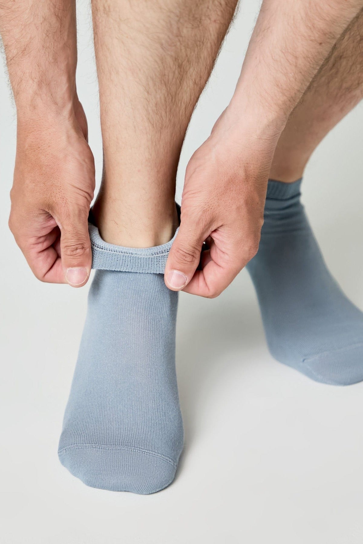 A person pulls on a pair of light blue ankle socks while standing against a neutral light gray background.