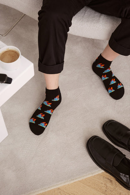 Black ankle socks featuring a repeating pattern of cartoon dragons are worn by a person sitting on a chair next to a white side table and a pair of black loafers on a light-colored rug.