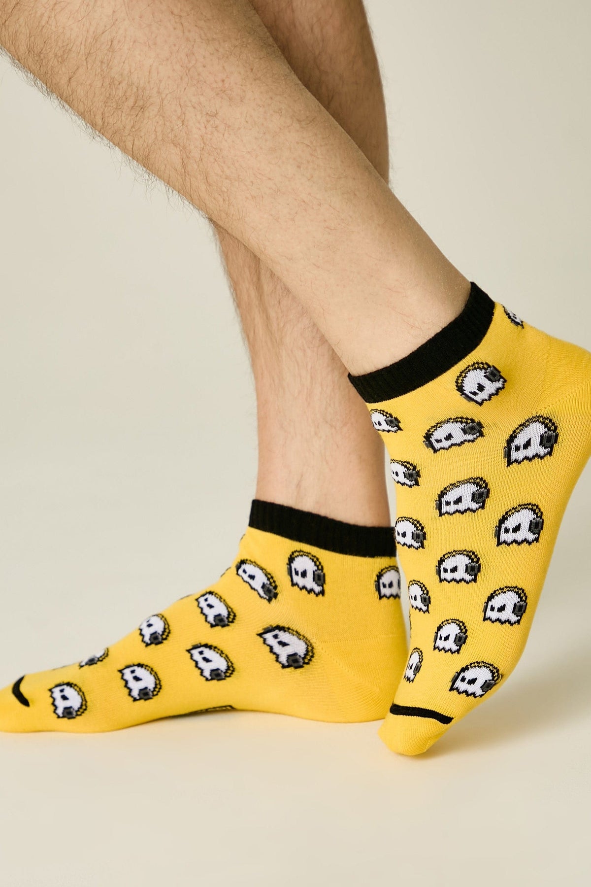 Ankle-length socks featuring a yellow base with a repeating pattern of white and grey pixelated ghost icons and a black ribbed trim worn on a person's feet.