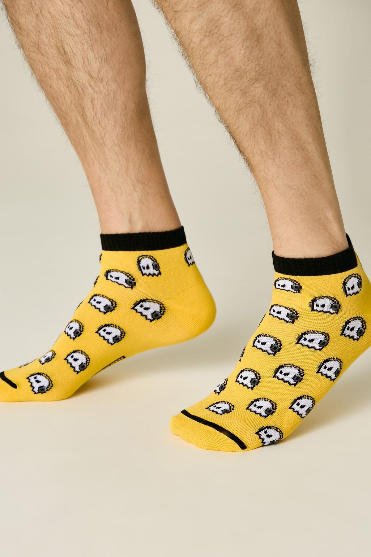 Ankle socks in bright yellow featuring an all-over repeating pixelated ghost pattern with black trim at the cuffs and toes, worn on a person's feet against a light background.