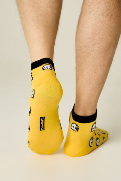A pair of bright yellow ankle socks with a black trim and a repeated white ghost pattern, worn on a person's feet against a light grey background.
