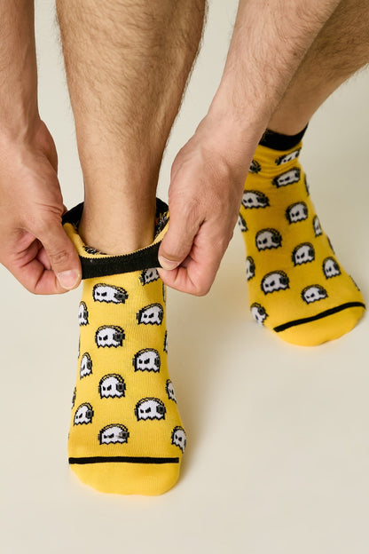 A person is putting on bright yellow crew-length socks featuring a repeating pattern of small, white, ghost-like arcade characters with dark outlines and black trim.