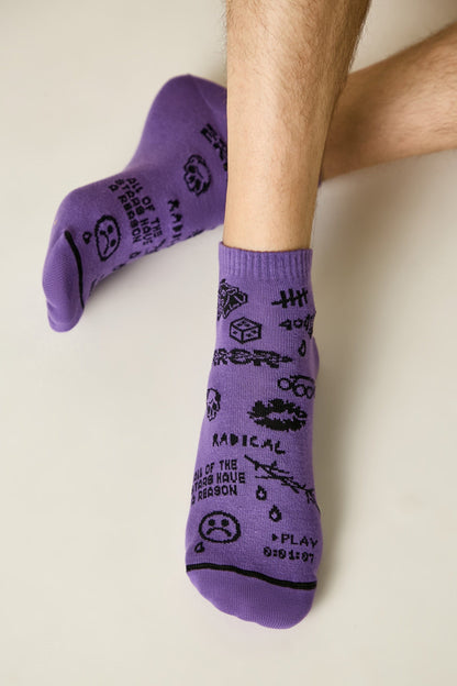 A pair of purple ankle socks feature various black printed graphic designs including text that reads "RADICAL" and "ALL OF THE STARS HAVE A REASON," a skull, a cube, a sad face, and playback icons.