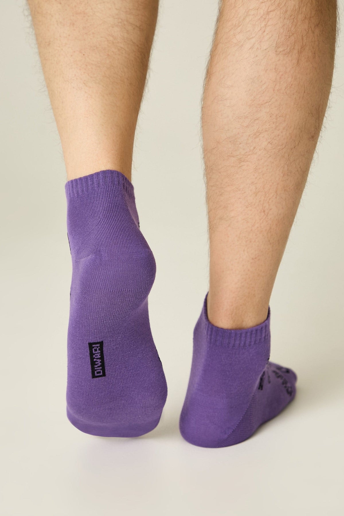 A pair of purple ankle socks worn on a person's feet against a plain light background, showing a small black brand label on the back of the left heel.