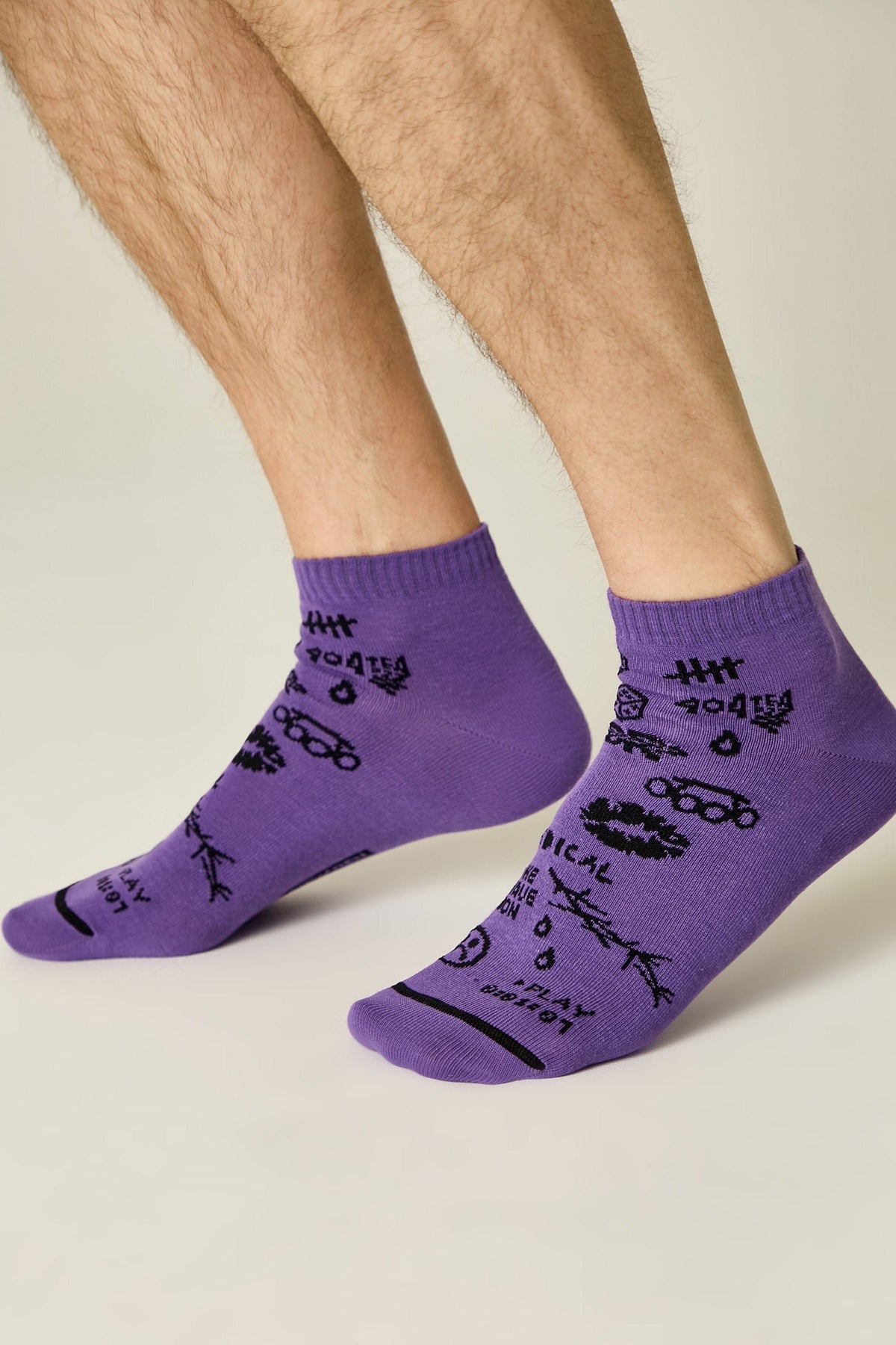 A pair of purple ankle socks with black illustrative patterns, including a smiley face, line drawings, and text accents, worn on a person's feet against a light background.