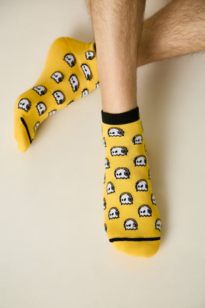 Yellow ankle socks feature a repeating pattern of white pixelated ghost characters and black trim at the cuffs and toes, worn on a person's feet against a light background.
