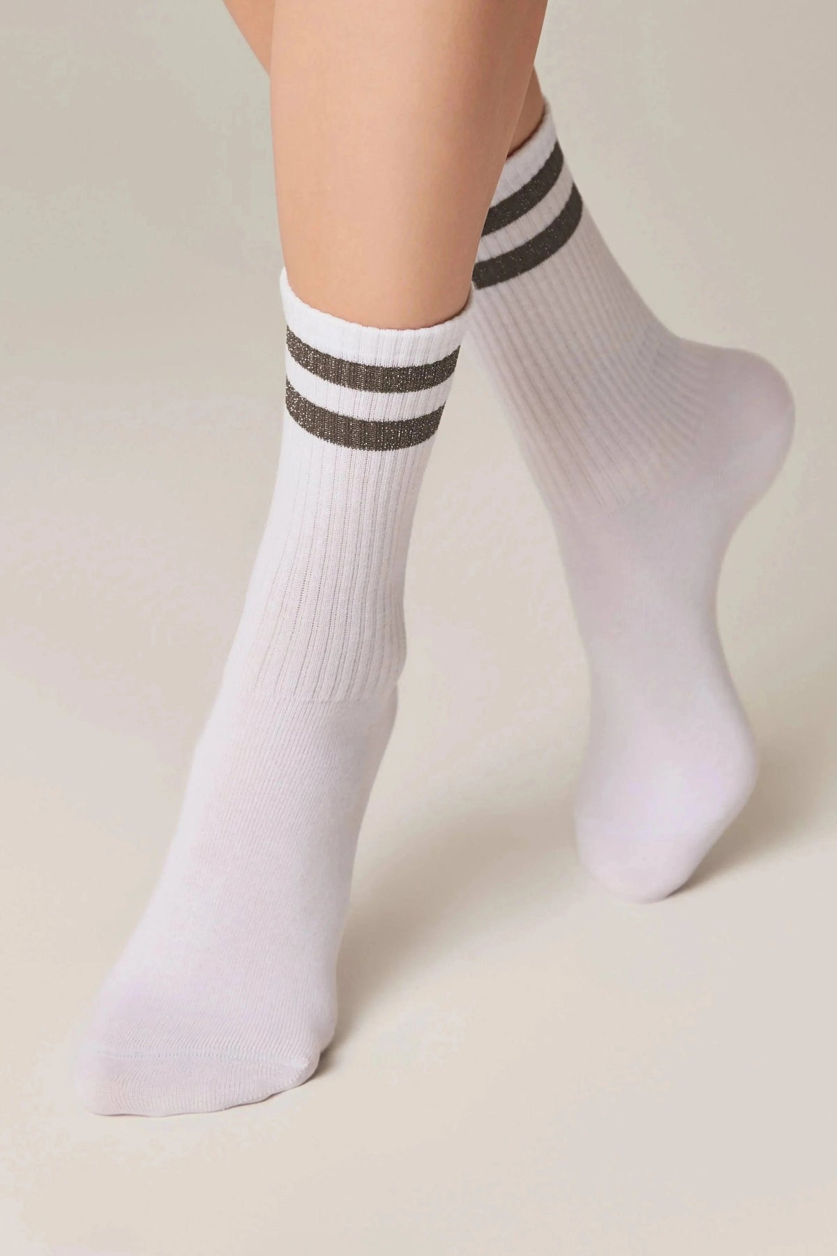 A pair of white crew-length socks featuring two horizontal glittery dark grey stripes around the ribbed cuffs, shown on a pair of legs.