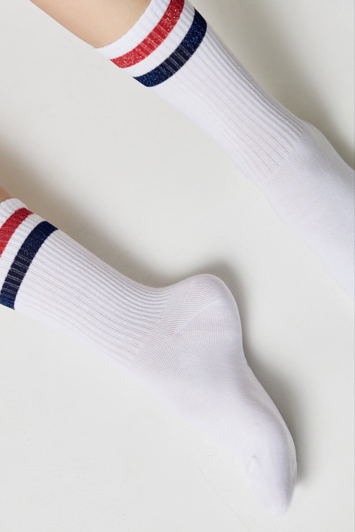A pair of white crew socks featuring two horizontal stripes in red and blue glitter near the top cuff, worn on a person's feet against a plain white background.