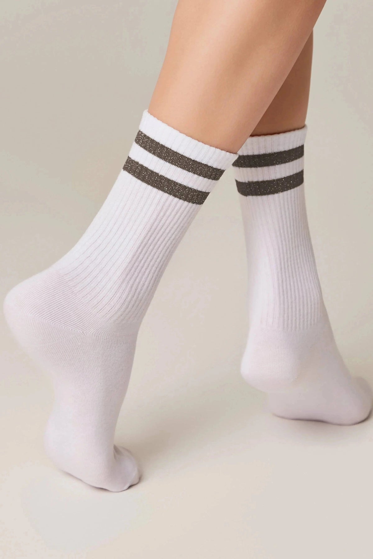 A pair of white crew-length ribbed socks featuring two dark glittery horizontal stripes on the upper cuffs, worn on a model against a plain light background.