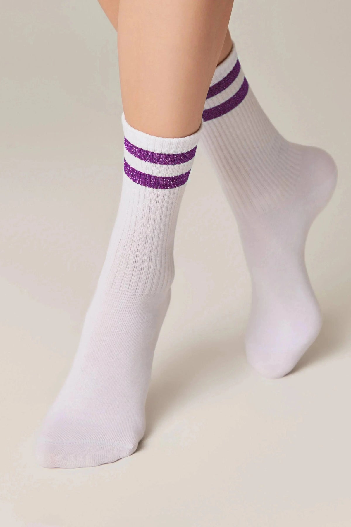 A pair of white crew-length socks featuring two horizontal purple glittery stripes near the top, worn on a person's feet against a plain light background.
