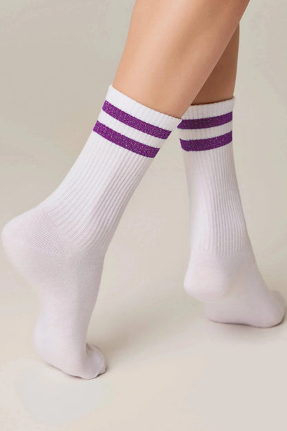 White crew socks with two purple glitter stripes around the ribbed cuff, worn on feet against a light background.
