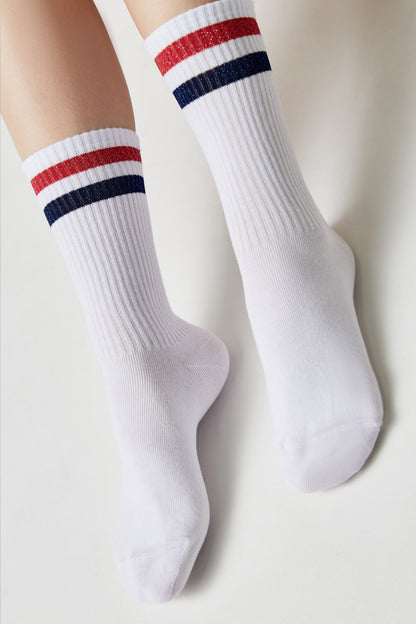 White crew socks worn on a person's feet, featuring a ribbed texture and two horizontal stripes in red glitter and navy blue near the top cuffs.