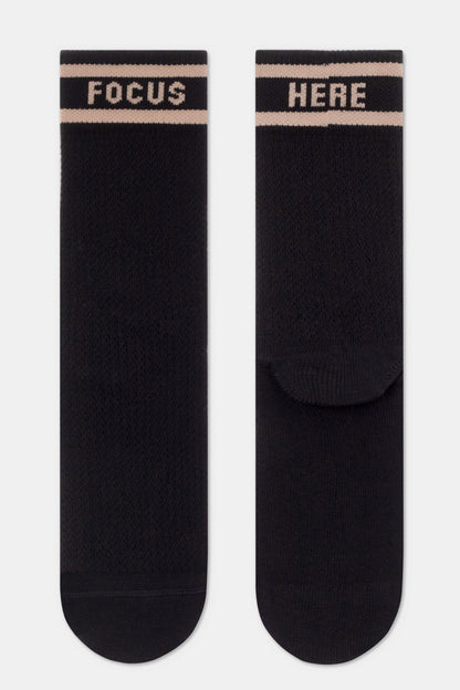 A pair of black crew-length socks featuring two horizontal light beige stripes near the top, with the word FOCUS printed on the left sock and the word HERE on the right sock in a matching light beige font.