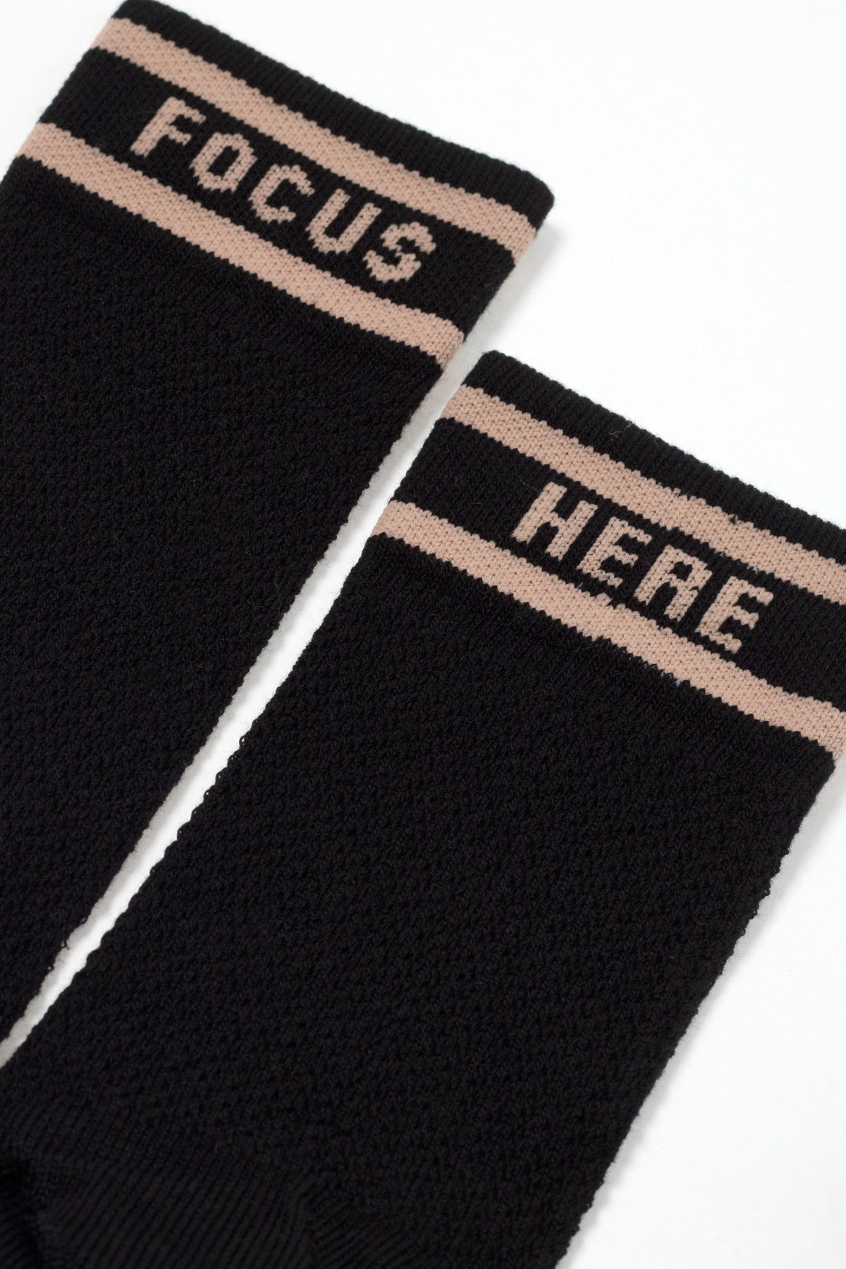 A pair of black crew-length socks featuring a tan stripe and the word FOCUS on one sock and the word HERE on the other in tan block lettering.