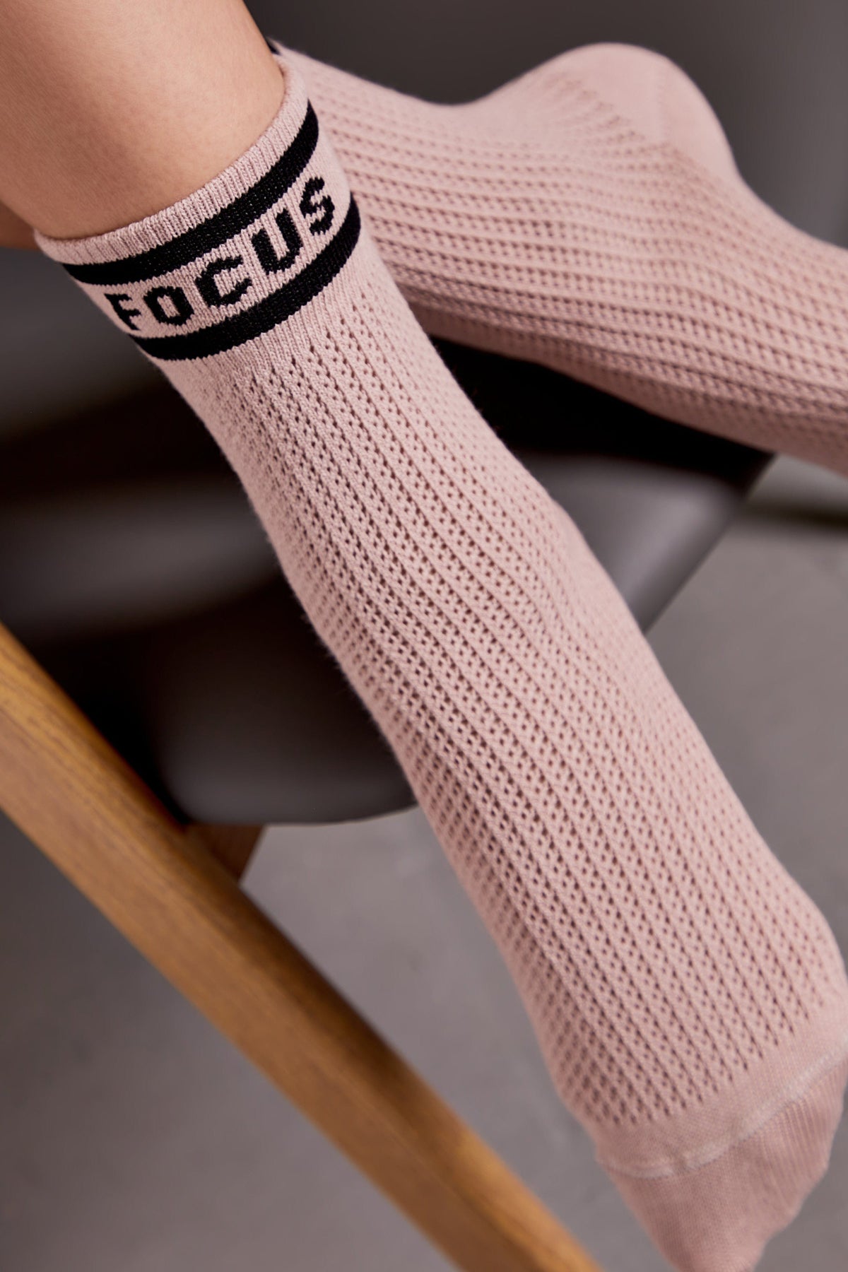 A pair of light pink, ribbed, mid-calf socks with the word FOCUS printed in black on a thick stripe at the top, worn by a person resting their feet on a grey chair.