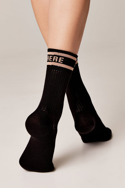 Black athletic crew socks with a beige ribbed cuff featuring the word HERE in bold lettering, worn on a person's feet against a neutral background.
