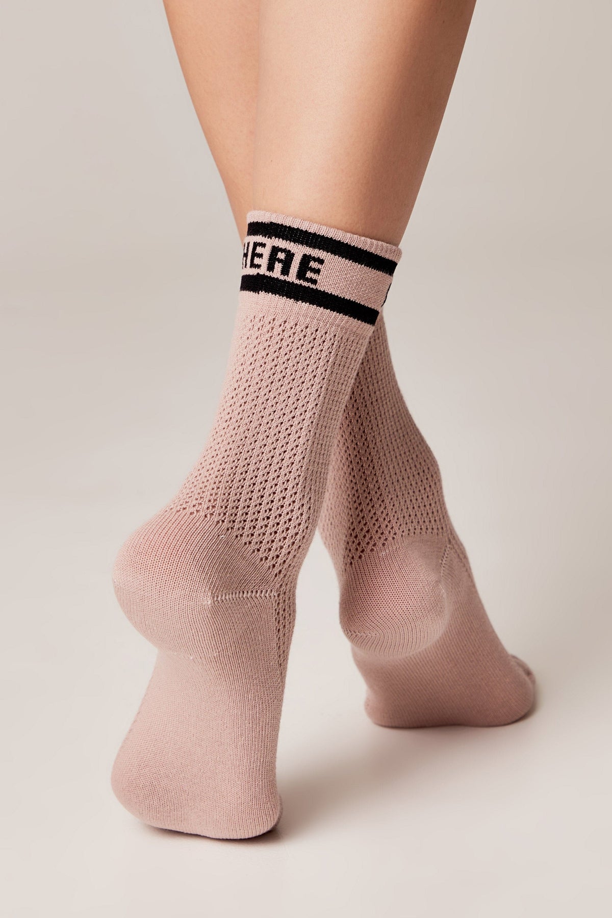 A pair of dusty pink crew socks featuring a breathable knit texture and black horizontal stripes with the word HERE printed in black on the cuff.