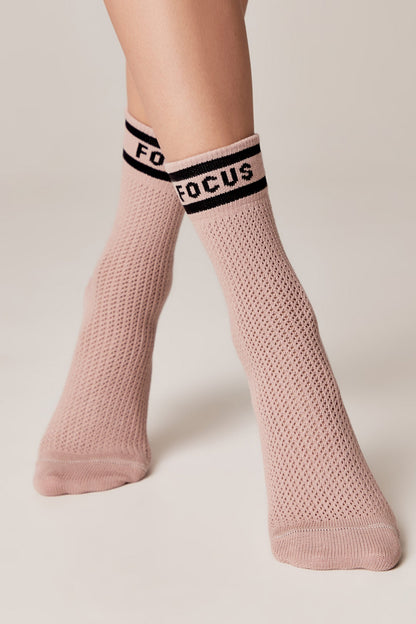 Light pink crew-length socks with a textured mesh knit pattern and a black ribbed cuff featuring the word FOCUS in bold white letters, worn on feet against a plain light background.