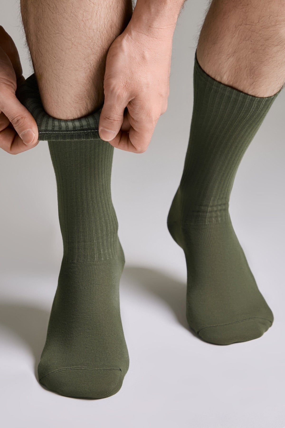 A person pulls up a pair of olive green crew-length socks on their lower legs against a plain grey background.
