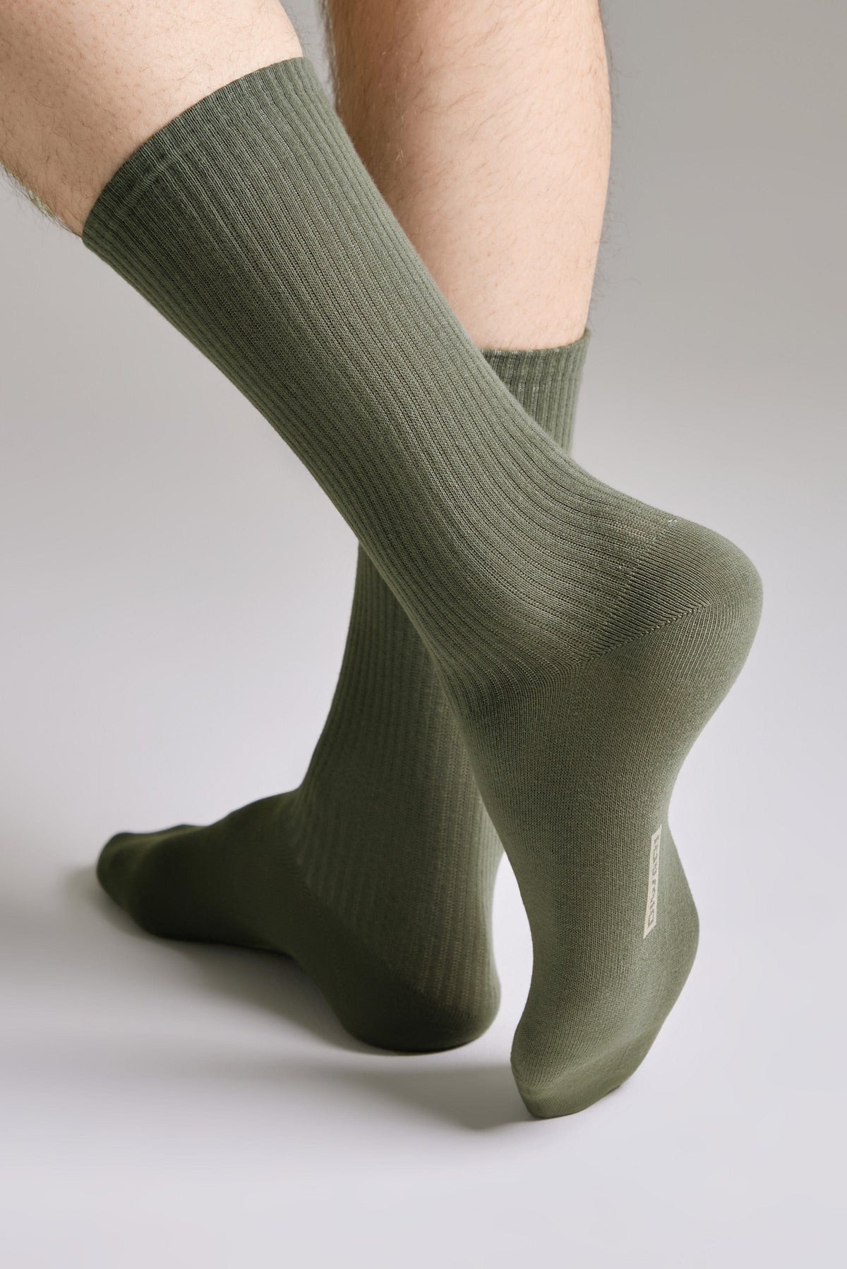 A pair of olive green ribbed crew socks worn on a person's legs against a plain grey background.