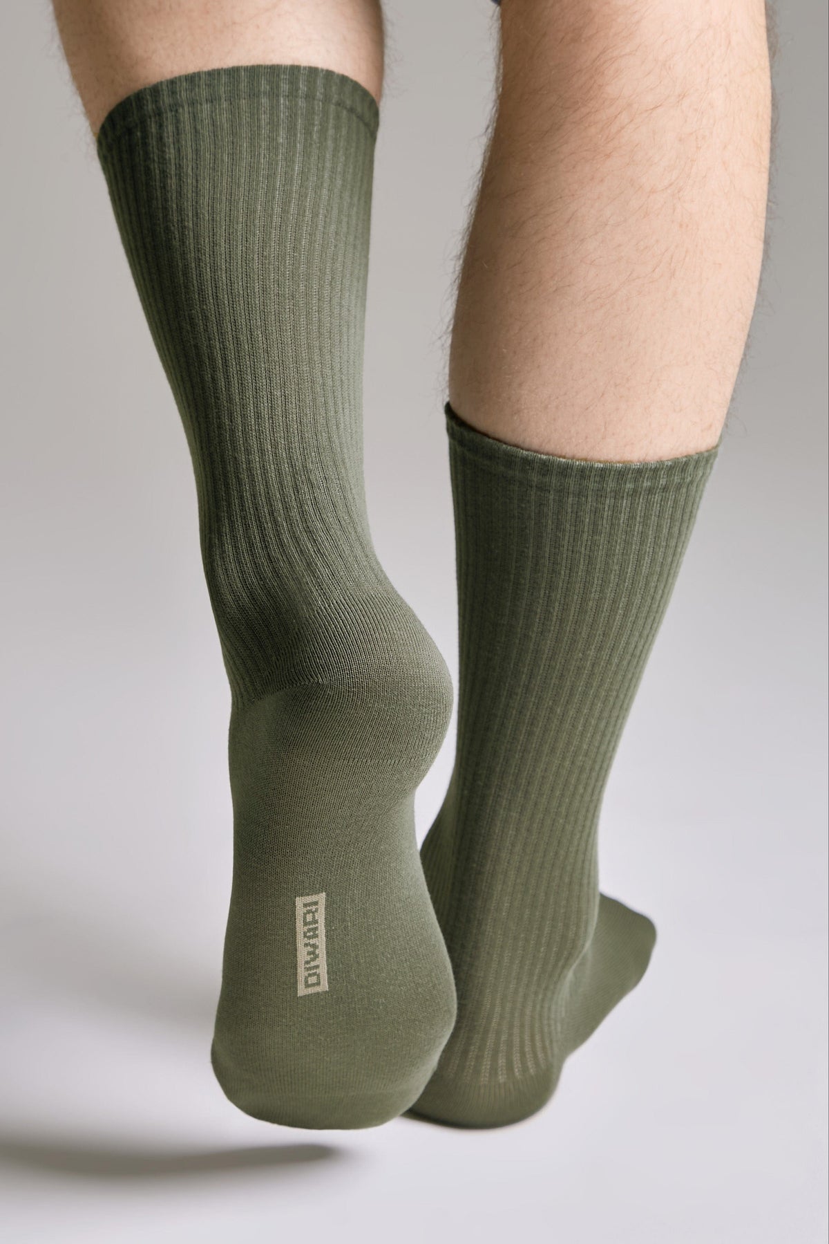 A pair of olive green ribbed crew socks worn on a person's feet, viewed from behind with a branding logo visible on the heel.