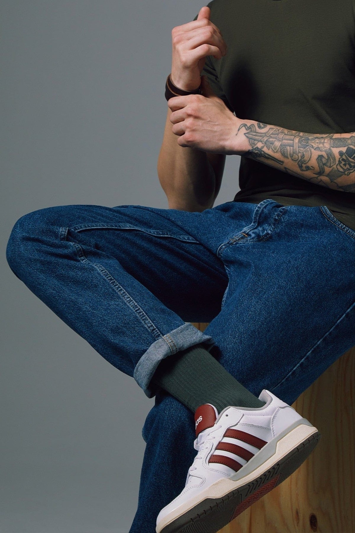 White sneakers with burgundy stripes and a brown tongue logo, worn with dark blue cuffed jeans and dark olive socks by a person with a forearm tattoo.