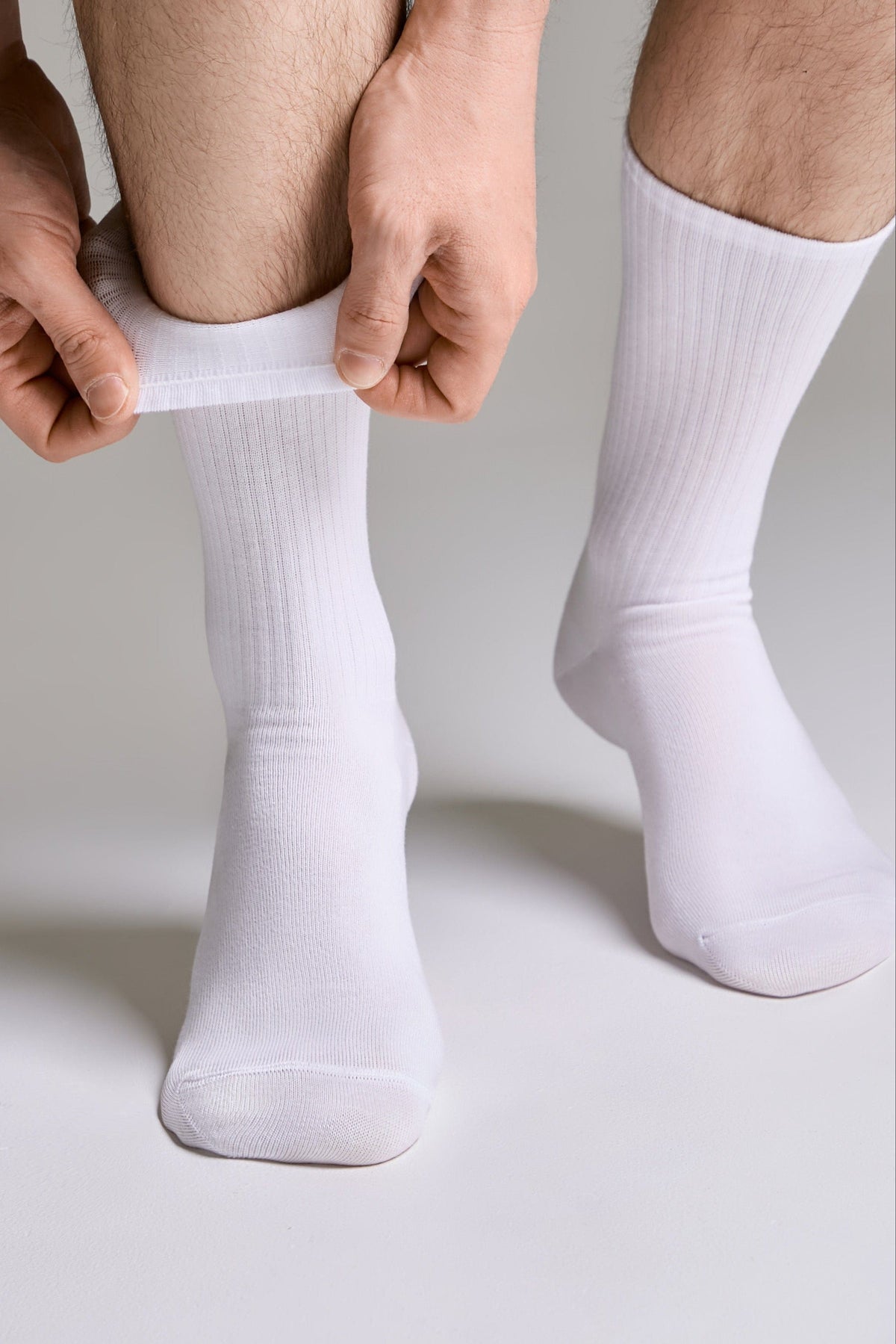 A person wearing plain white crew socks on their feet while pulling up the cuff of the left sock against a solid grey background.