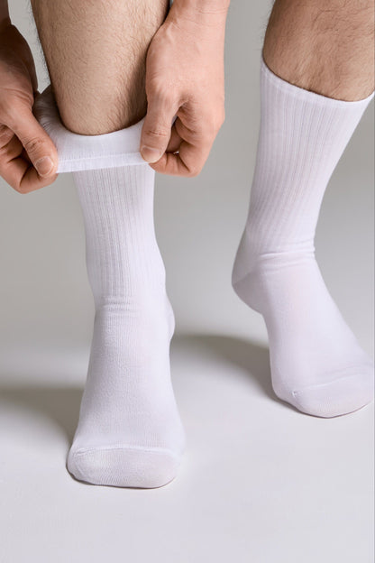 A person wearing plain white crew socks on their feet while pulling up the cuff of the left sock against a solid grey background.