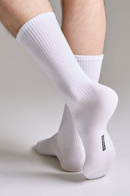 A pair of ribbed white crew socks worn on a person's feet against a neutral grey background, with a small rectangular black logo visible on the heel.