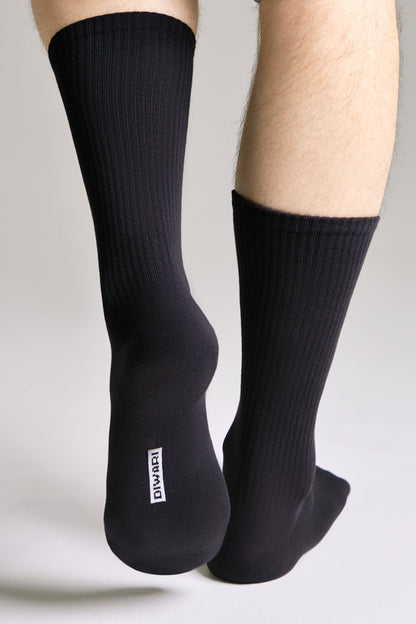 A pair of black ribbed crew-length socks worn on feet, featuring a white DIWARI brand label on the underside of one heel.