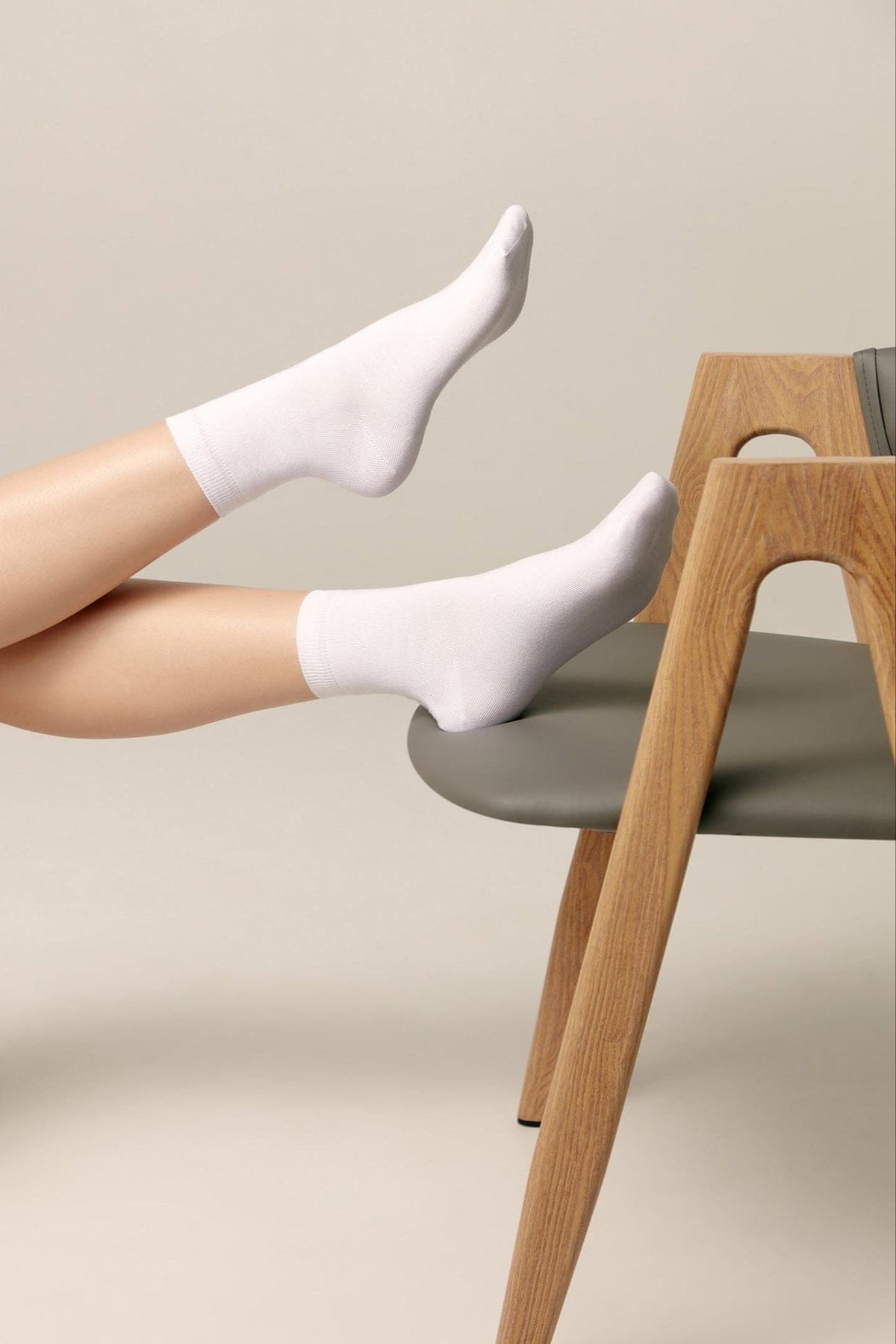 White ankle socks worn on a person's feet resting on a gray chair with wooden legs.