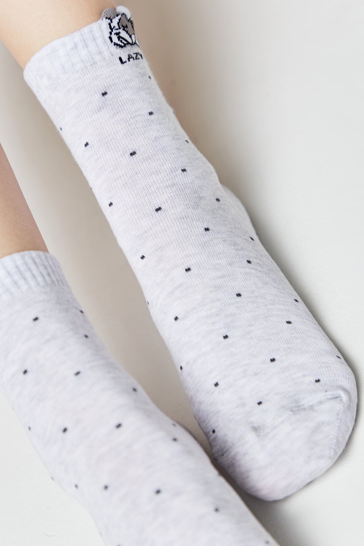 A pair of light grey ankle socks featuring a subtle pattern of tiny dark dots and a small cartoon character embroidered at the top of the cuff with the word lazy below it.