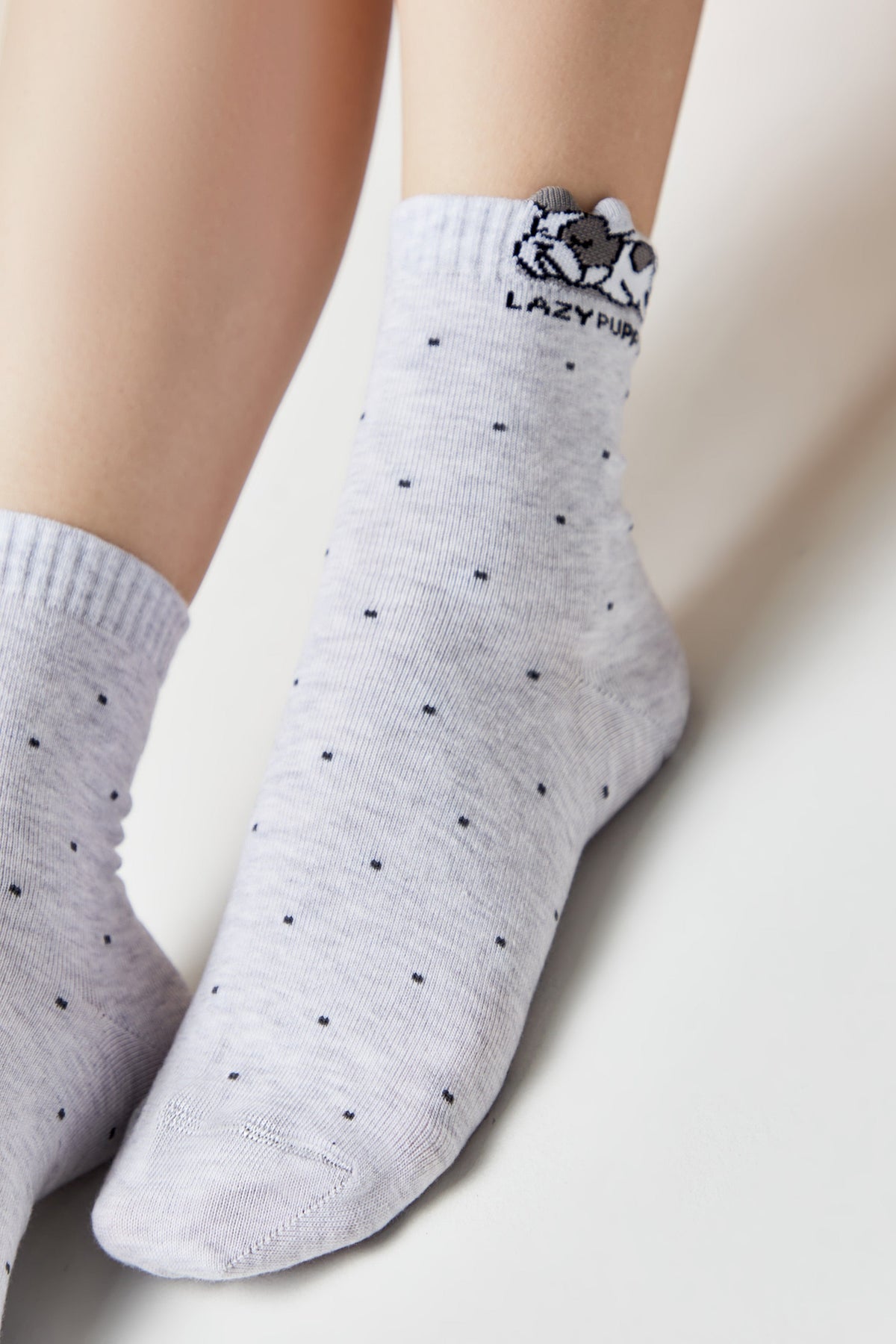 A light grey ankle sock with a small dark dog face graphic and the words LAZY PUP printed on the cuff, featuring a subtle black polka dot pattern across the fabric.
