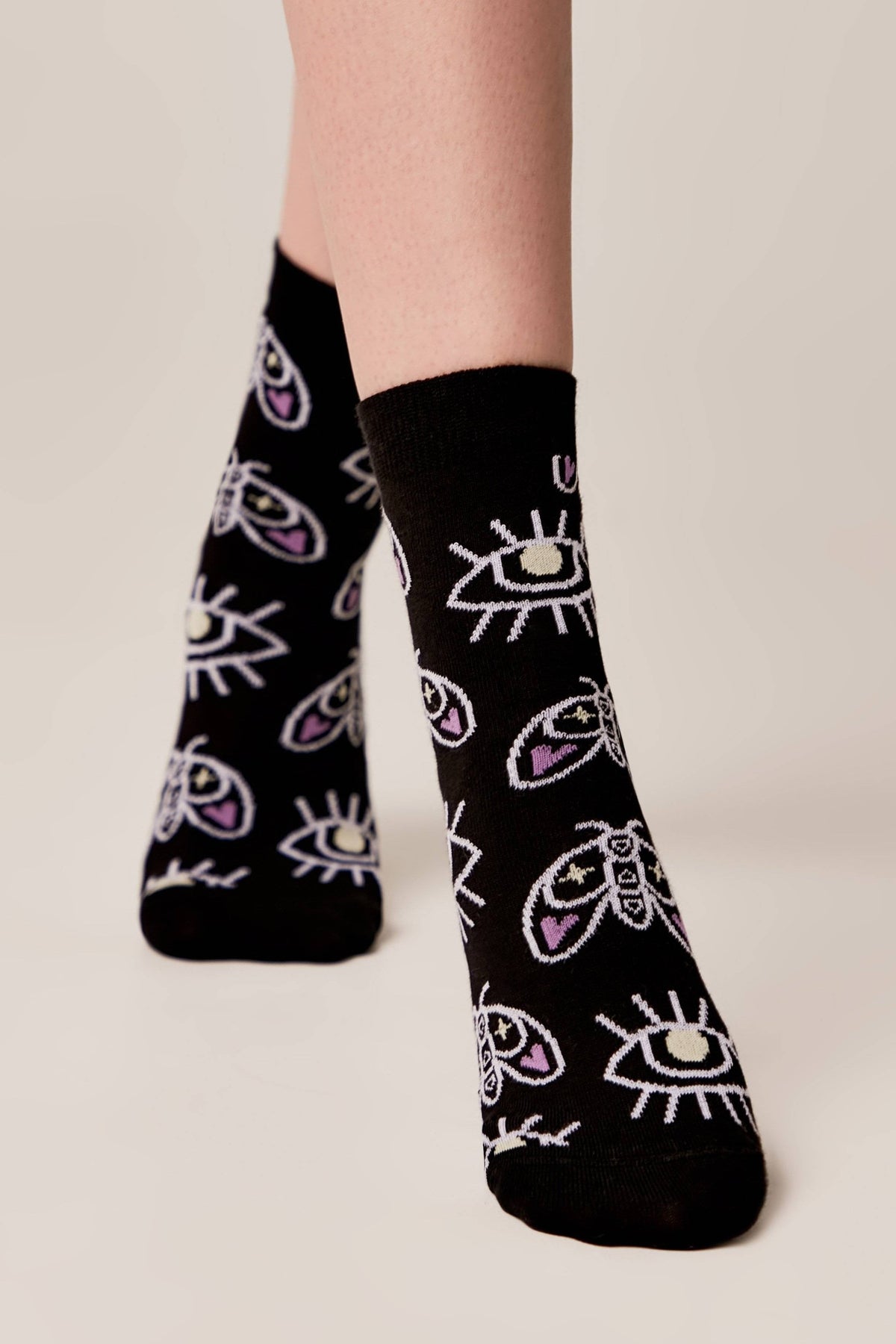 Black socks featuring a repeating pattern of white and purple moths and stylized eyes worn on a person's feet against a neutral background.