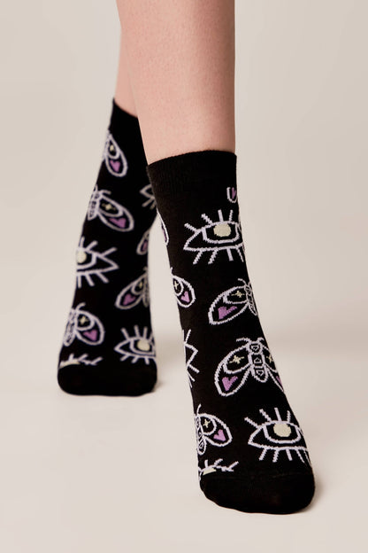 Black socks featuring a repeating pattern of white and purple moths and stylized eyes worn on a person's feet against a neutral background.