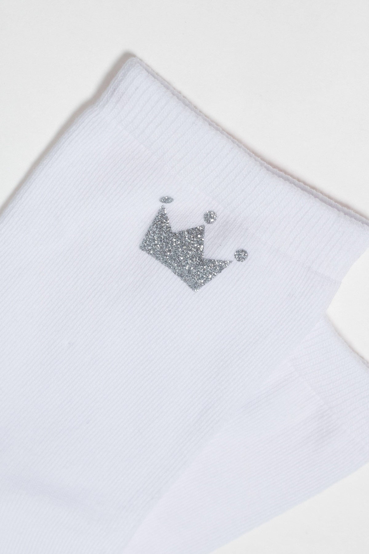 A white ribbed ankle sock features a small, sparkly silver crown icon embroidered on the side.