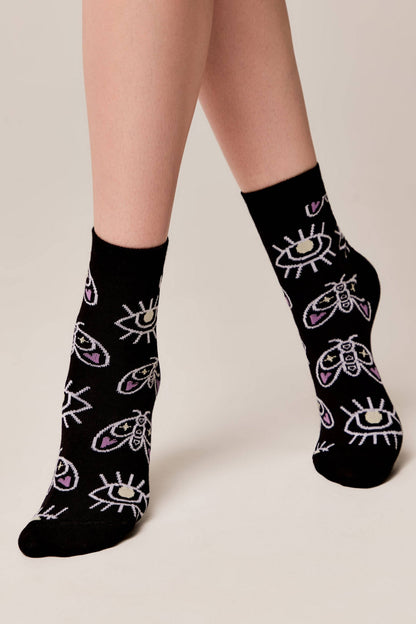 A pair of black ankle socks featuring a repeating pattern of white and purple eyes and moth illustrations worn on a person's legs against a light beige background.