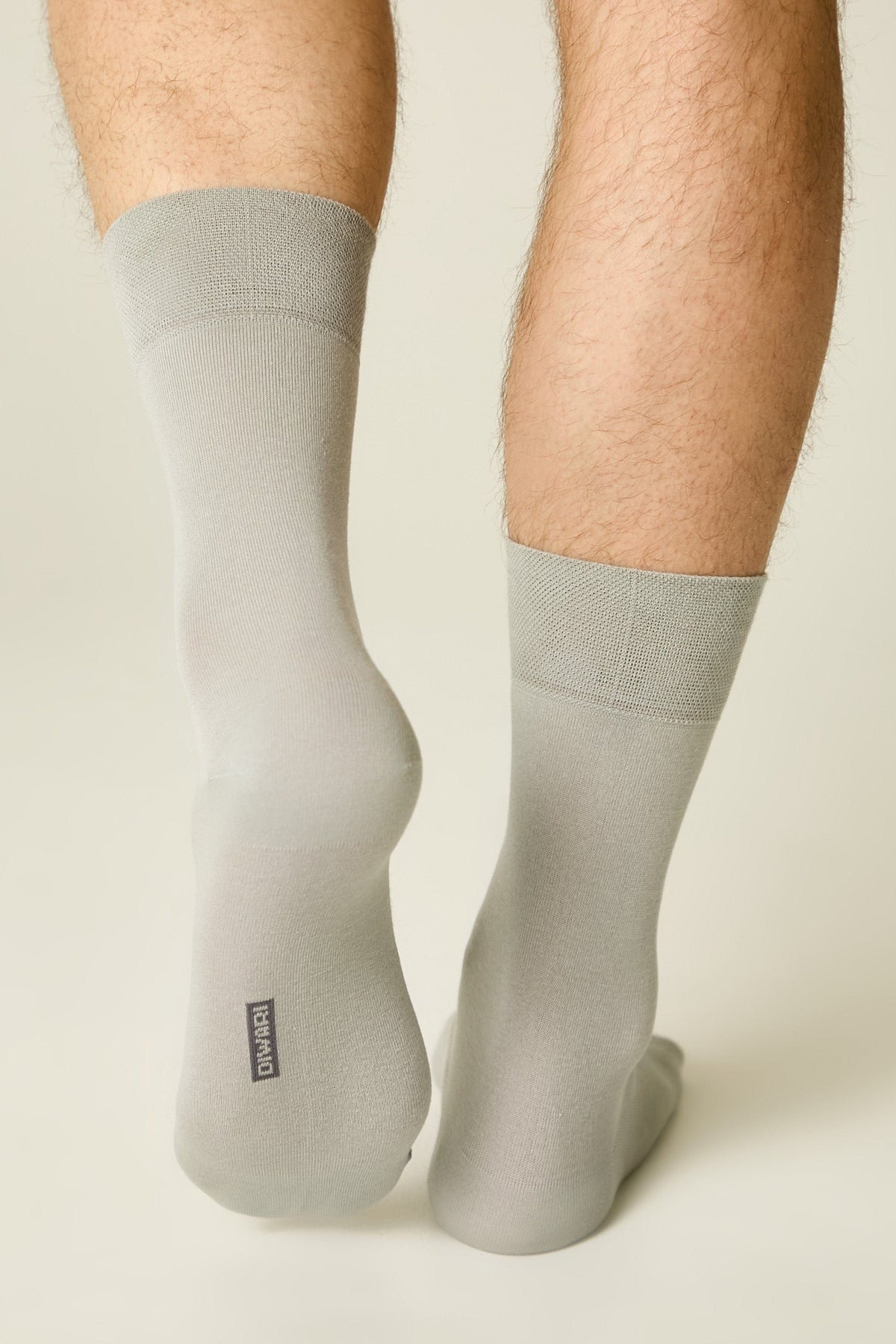 A person wearing a pair of light grey mid-calf socks with a small black rectangular logo on the heel of the left sock.