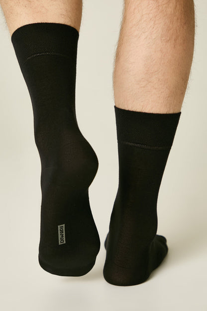 A pair of black crew socks worn on a person's lower legs, showing a small grey rectangular logo on the heel of one sock against a light grey background.