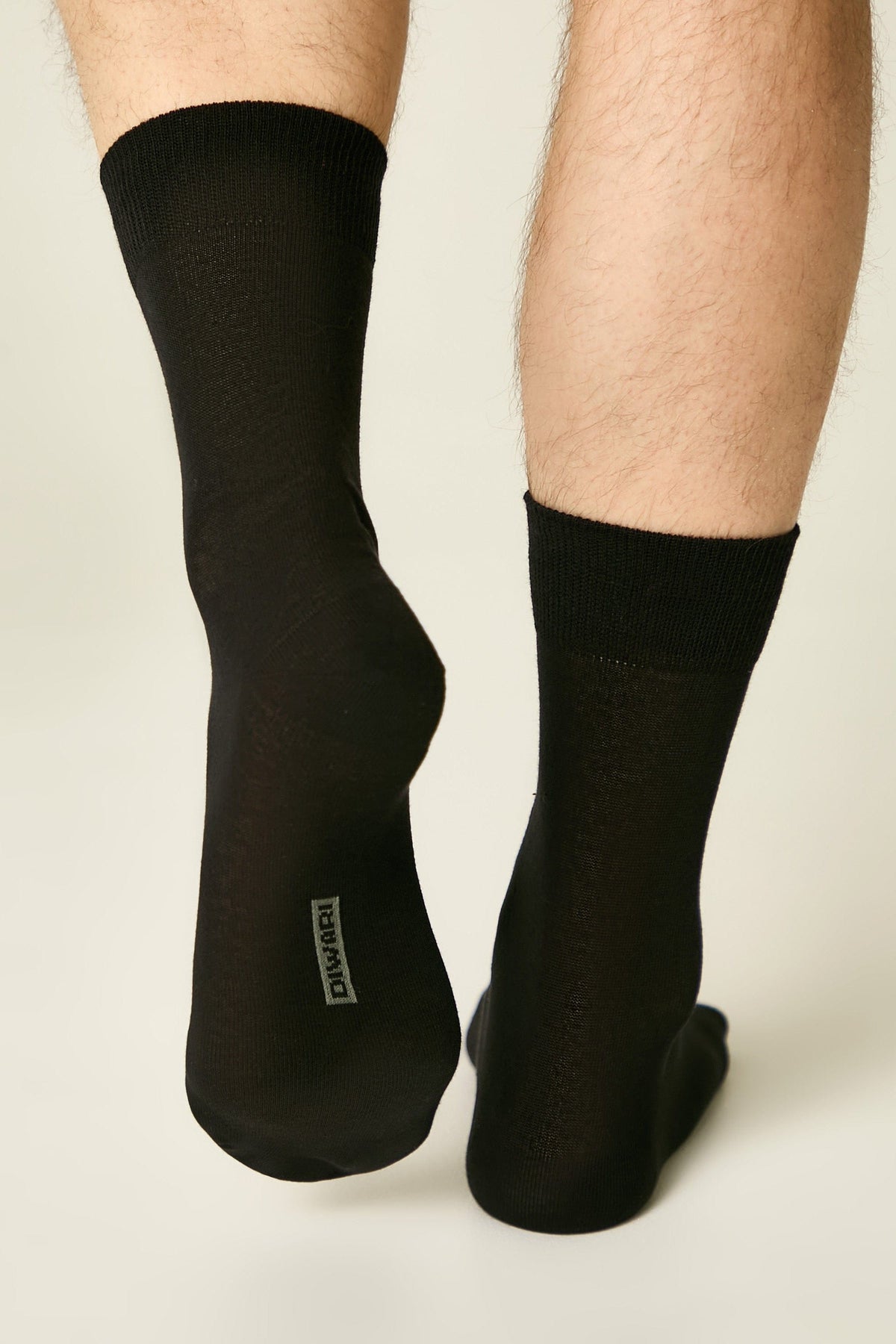 A pair of black crew-length socks worn on a person's feet against a plain light background, with a small grey brand label visible on the bottom of the left heel.