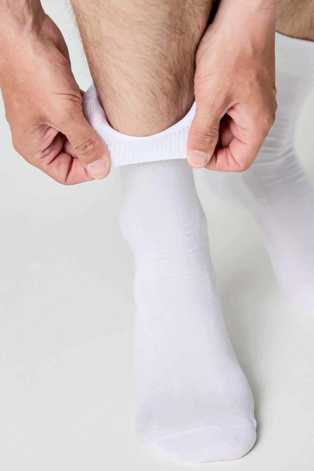 A person pulls up a pair of clean white crew socks on their lower leg against a plain white background.
