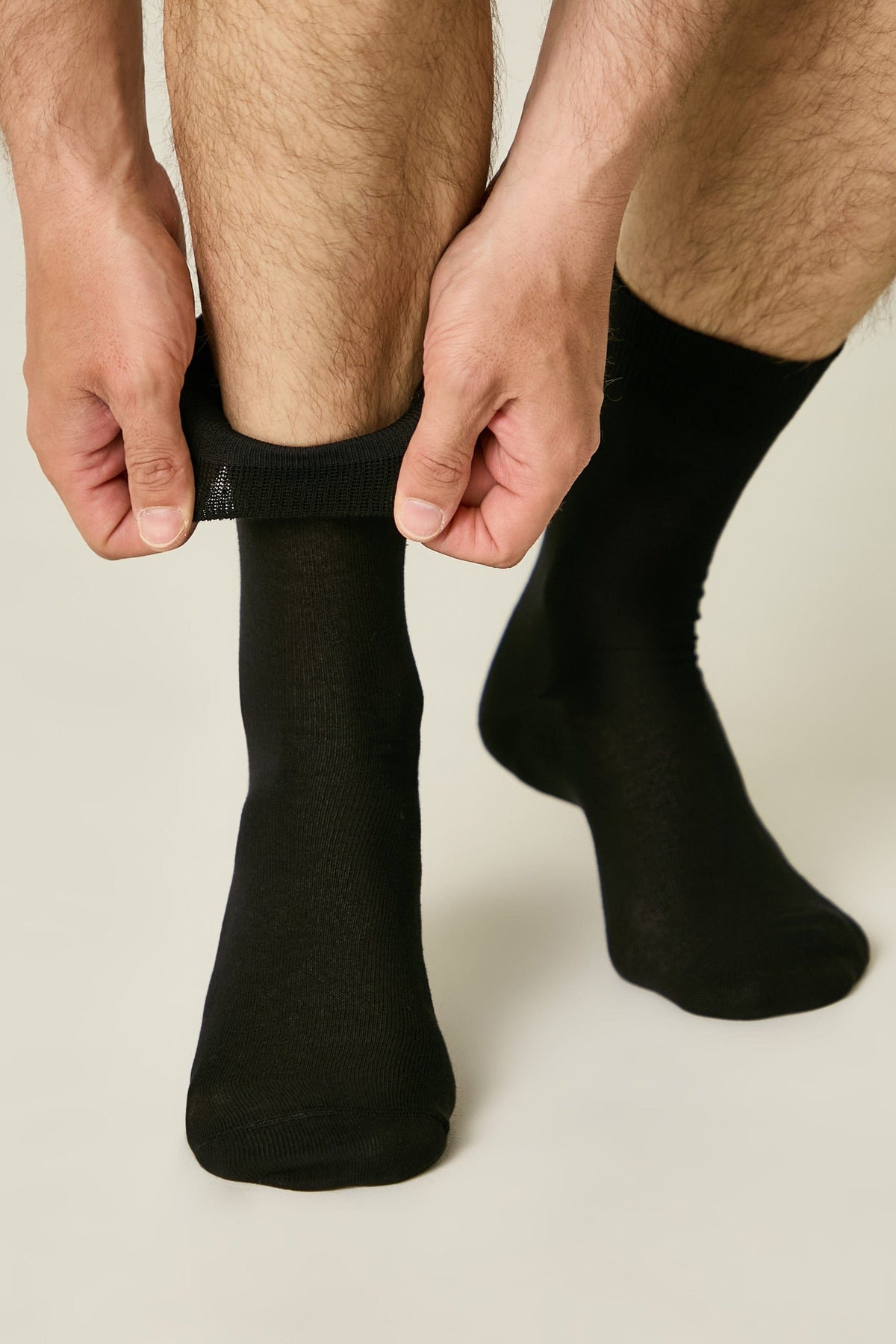 A pair of plain black mid-calf socks being put on by a person, showing the elastic cuff held by hands against a light background.