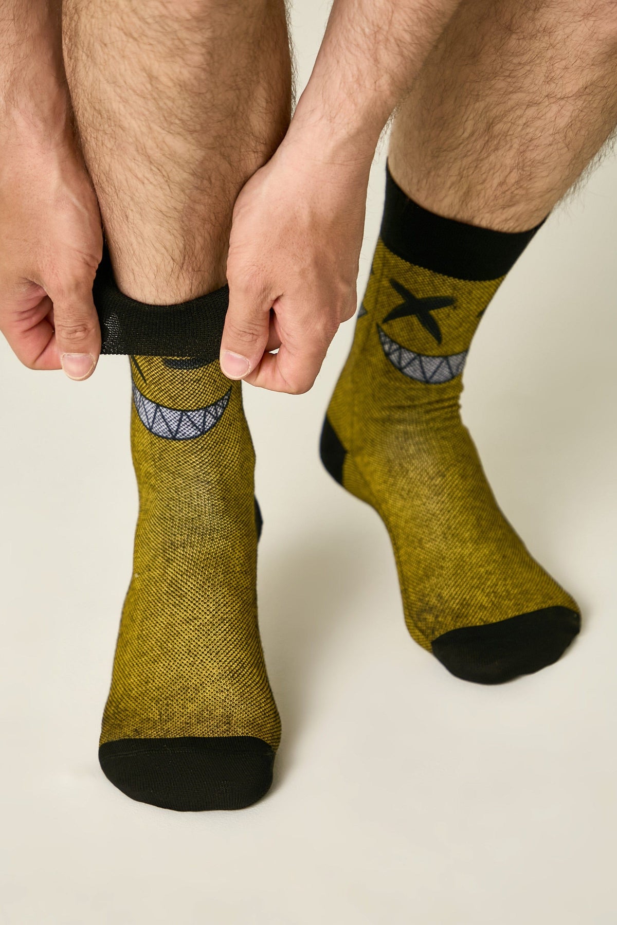 Crew socks in a mustard yellow textured knit feature a black cuff, black toe, and a graphic of a grinning mouth with X-shaped eyes worn on a person's feet.
