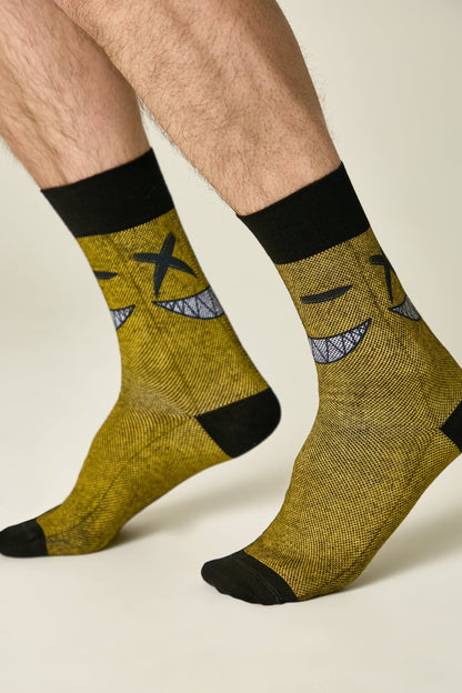 Crew socks in a mustard yellow textured knit feature a black graphic of a mischievous smiling face with X-shaped eyes and black cuffs and toe caps.
