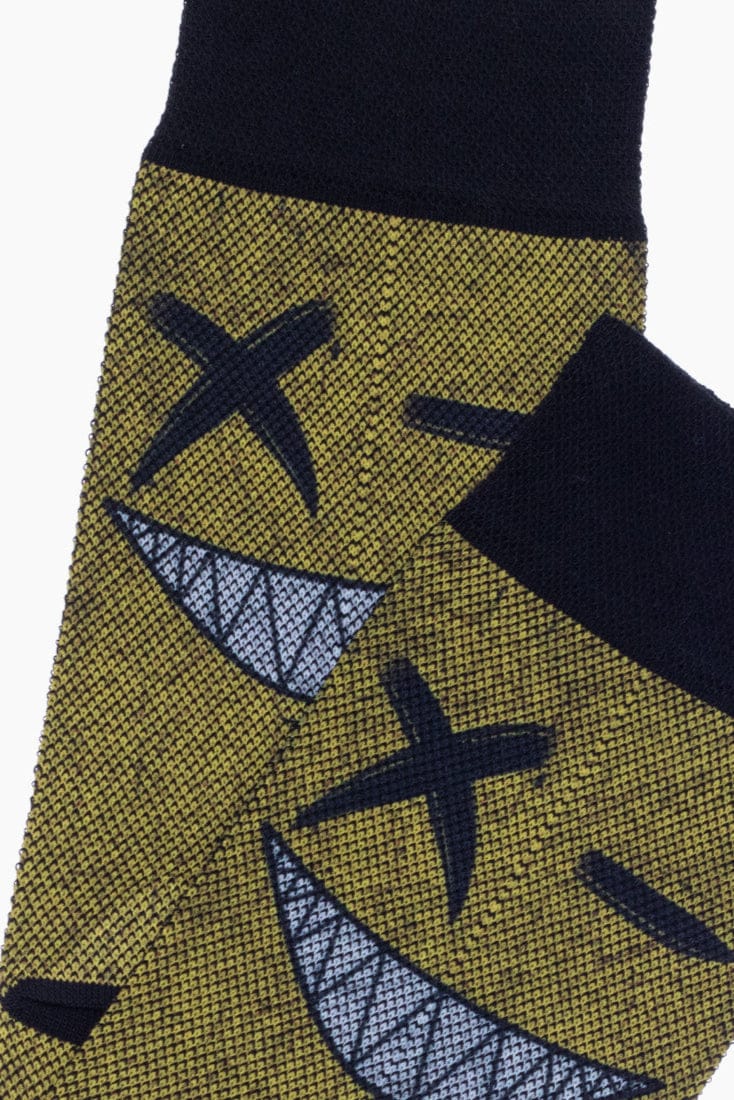 A pair of crew socks featuring a dark yellow fabric with a black knit pattern and a graphic print of a cartoon face with x-shaped eyes and a wide, sharp-toothed grin.
