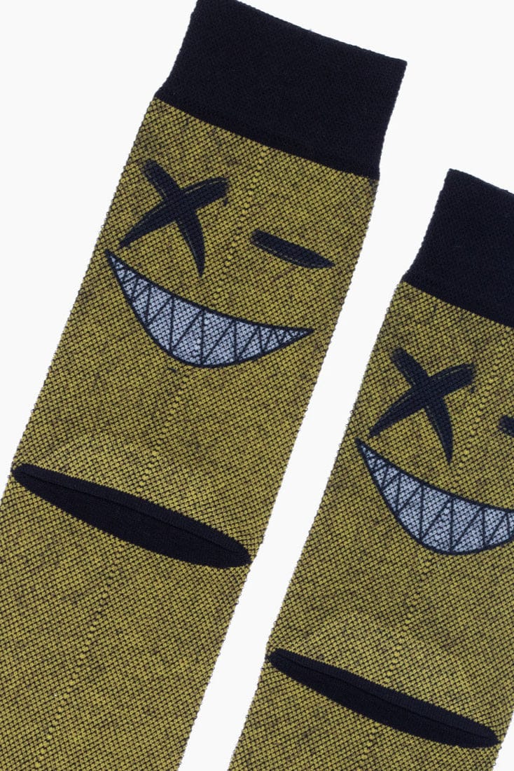 A pair of olive green crew socks featuring a dark embroidered design of a smiling face with an x-shaped eye, a winking eye, and sharp teeth, finished with solid black cuffs.