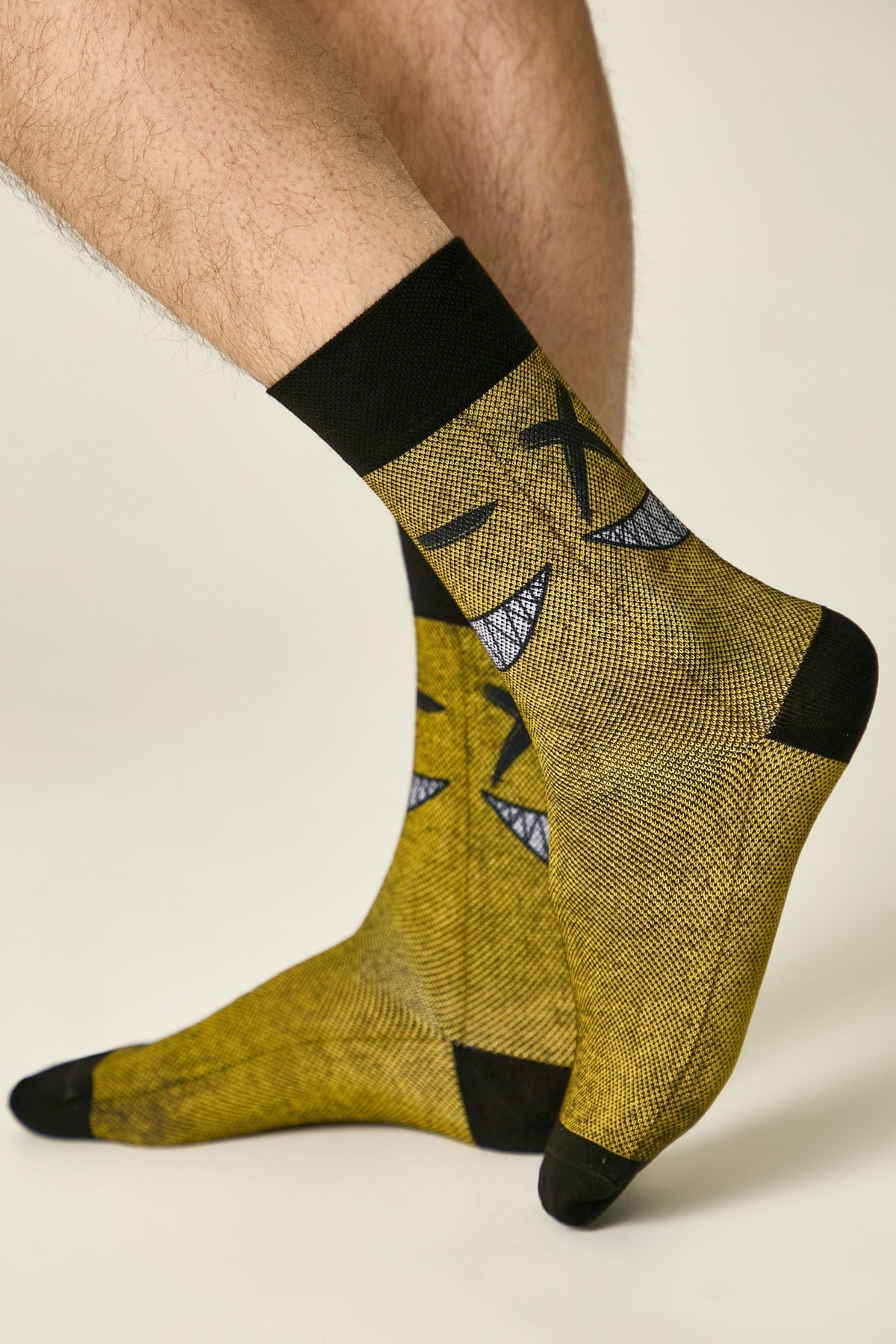 Crew socks in a mustard yellow fabric feature a repeated black graphic pattern of a stylized character with X-shaped eyes and a wide, toothy grin on a person's legs.