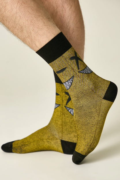 Crew socks in a mustard yellow fabric feature a repeated black graphic pattern of a stylized character with X-shaped eyes and a wide, toothy grin on a person's legs.