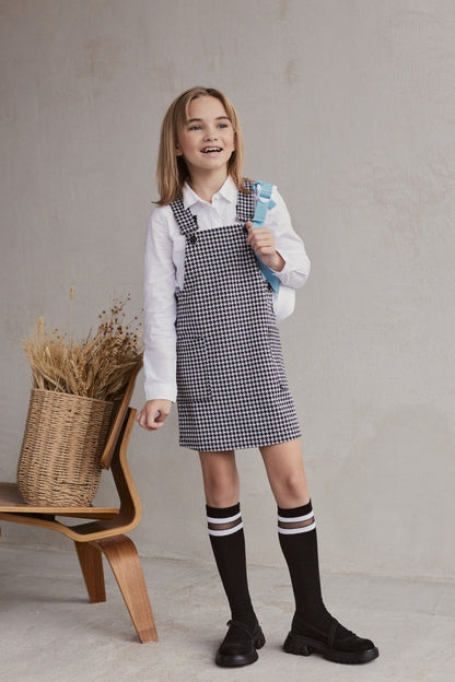 A young girl stands wearing a white long-sleeved collared shirt under a black and white houndstooth pinafore dress, paired with black knee-high socks featuring white stripes and black platform shoes, standing next to a chair holding a woven